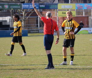 Scheidsrechter Arthur Ebink heeft gefloten nadat de VELO-doelman de bal te lang vasthield. Phil Altimari hoopt op een kans.